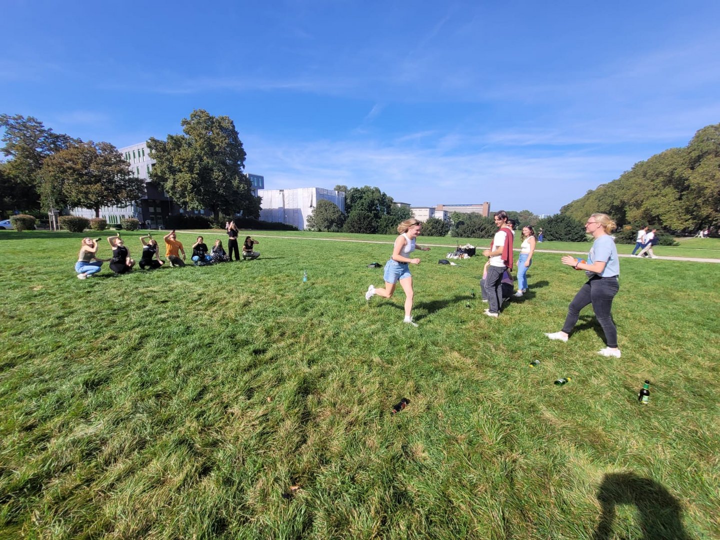 The picture shows a bunch of people in a parc. On the left, some people are kneeling on the ground and drinking from bottles. In the middle you can see one person running towards another group of people on the right.